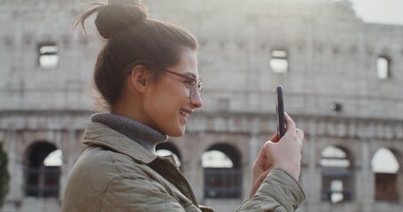 A warmly dressed young woman in glasses smiles while taking photos of the sights of Rome, standing on an observation deck near the Colosseum. Video shooting from the side. Video in 4k, red komodo. © m-art