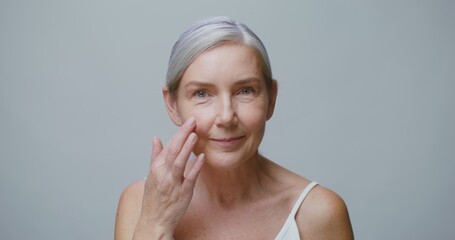 An adult model of European appearance with gray hair pulled back in a tail applies moisturizer to...