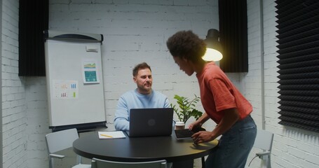 A young man of European appearance working at a laptop, welcomes a young woman who comes into his...