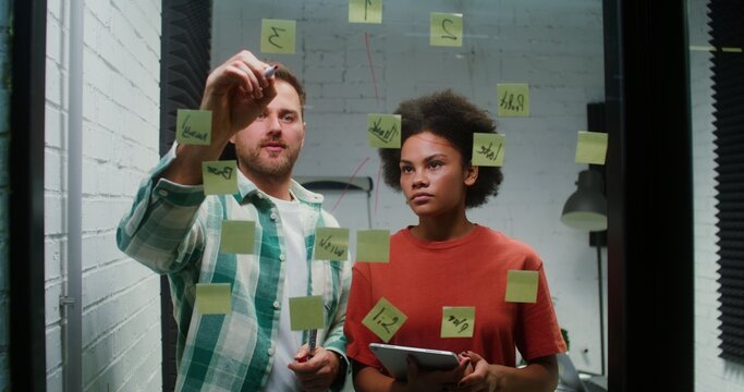 A Caucasian man and an African American woman are planning, drawing a diagram of work on a glass board with stickers and using a tablet during a working day in the office. Brainstorm, business concept