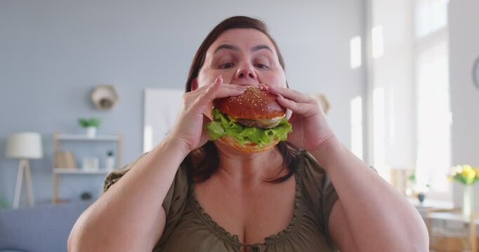 Close up of smiling overweight fat woman enjoying big burger at home. Cheerful plus size female indulging in fast food, showing carefree attitude, love for tasty meal and unhealthy eating habits.