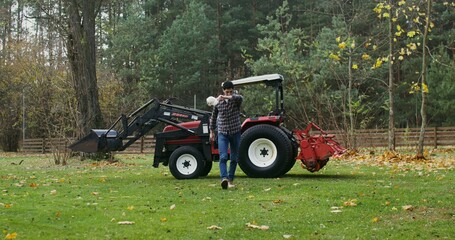 Young dark-haired man climbs out of an agricultural vehicle tired after working in the field, taking off his baseball cap and wiping his forehead with his hand, on a sunny autumn day