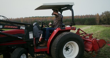 Young black haired male farmer eating a sandwich and drinking tea from a metal mug, while sitting in the open cabin of a modern agricultural tractor parked in the middle of a field for lunch