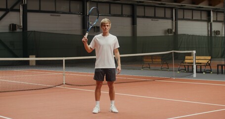 Obraz premium A young male tennis player of European appearance with a serious face throws a tennis racket behind his head, looking directly at the camera, standing on an indoor tennis court