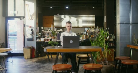 Young beautiful woman with a very short haircut, wearing glasses, works remotely in a stylish coffee shop, typing on a laptop while sitting at the table and drinking coffee
