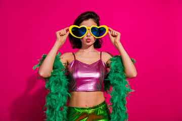 Stylish woman in shiny top with heart sunglasses posing against vivid pink background in playful retro-themed studio setup