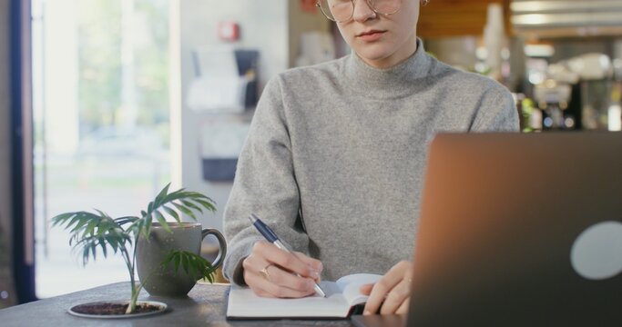 Online learning at the table in a modern coffee shop. A girl with very short hair makes notes with a pen in a notebook while sitting in front of an open laptop