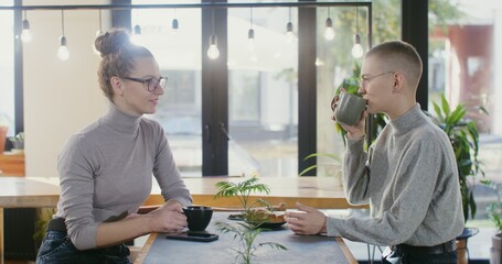 Two beautiful young women of European appearance communicate with each other, laughing and rejoicing, sitting opposite each other in a cozy stylish coffee shop over a mug of coffee