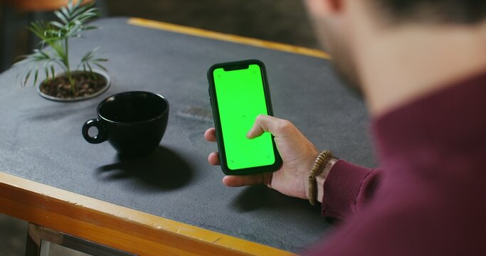 A young dark-haired man uses a green screen phone while sitting at a desk. Filming from behind, the man's face is not visible. Copy space, chroma key