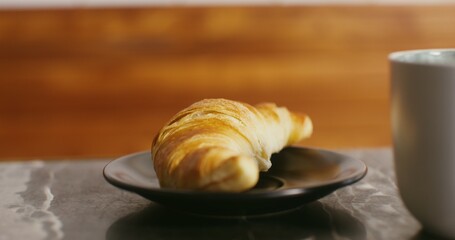 An appetizing fresh croissant lies on a small black plate on the table next to a coffee mug, close-up