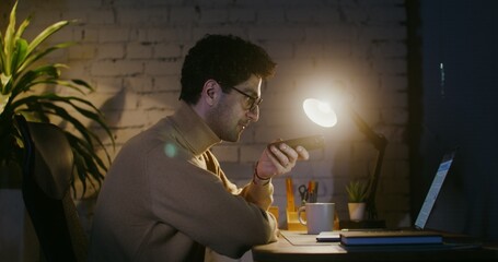 Young man in glasses with stubble on his face talking hands-free on a mobile phone while sitting at a table in front of an open laptop in the office, under the light of a lamp