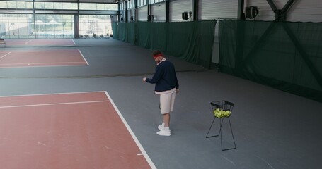 A young man of European appearance takes a tennis ball from a basket full of balls and throws it over the tennis net, hitting the racket, playing in an empty indoor tennis court