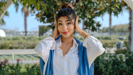 Naklejka premium A young brunette businesswoman in a tree-filled park on a spring day, looking at the camera while covering her ears with her hands to make the 