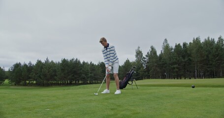 Obraz premium A young man of European appearance, dressed in white clothes, hits a ball with a golf club and peers into the distance, playing golf on a beautiful lawn