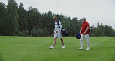 Two men of European appearance with a large age difference walk the golf course with bags of clubs...