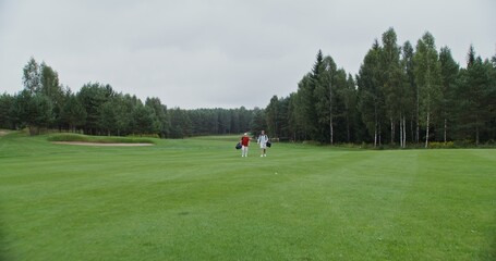Fototapeta premium father and son walk along the golf course, carrying golf clubs in their bags. Two men of different ages are going to play golf on a large well-groomed field surrounded by dense green forest