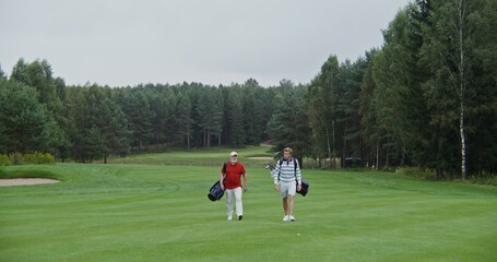 Two men carry golf gear in bags on their backs, walking on a neatly cropped playground and talking to each other. Business meeting on the golf course during the game