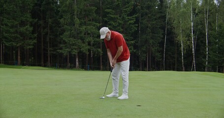 An elderly man with a gray beard, dressed in sportswear, plays golf, hitting a golf ball with a club. Unsuccessful attempt to drive a golf ball into the hole, missed and lost