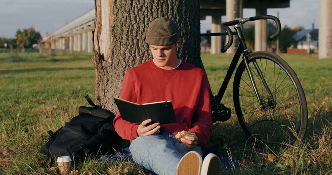 A young man of European appearance in a knitted hat reads a book and eats a sandwich, sitting on the ground under a tree next to the bridge. The bike stands under the tree next to the guy