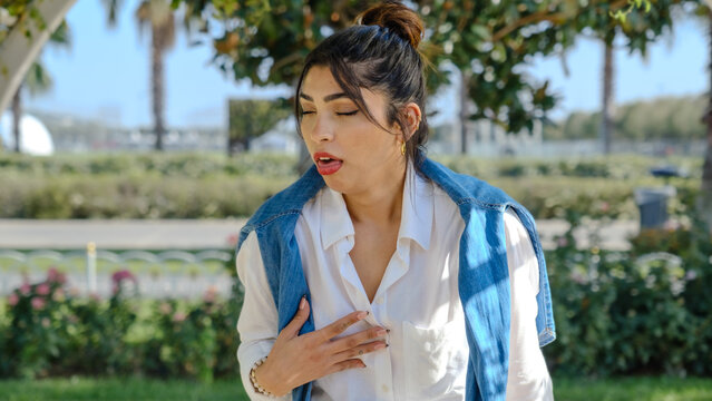 A young brunette businesswoman in a tree-filled park on a spring day, looking at the camera with facial expressions that indicate she is feeling nauseous