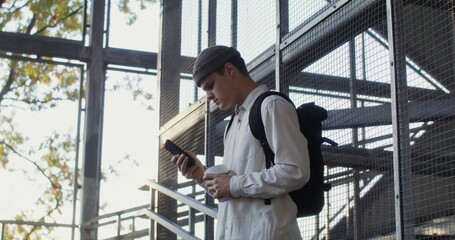 A young man of European appearance in a knitted hat uses a mobile phone, and drinks coffee while standing on the steps of a metal staircase
