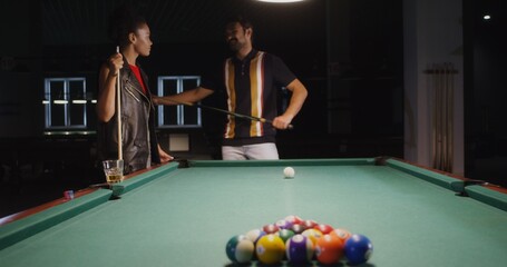 Fototapeta premium A young man explains and shows a young African American woman how to hold a cue and break a triangle in billiards. Fun pastime with talking and playing