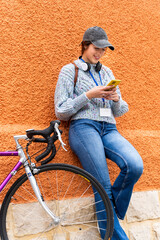 Fototapeta premium Young woman using smartphone while leaning on orange wall with bicycle nearby
