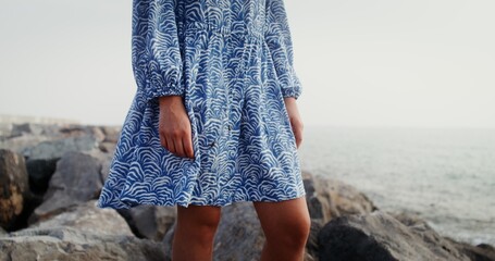 The wind develops the hem of the dress of a young tanned woman standing on a rocky seashore. No...