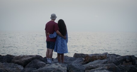 A young couple of lovers stand embracing on the seashore and look into the distance. Rocky shore, video filming from the back, no faces visible, evening, dusk. 4k video, red komodo