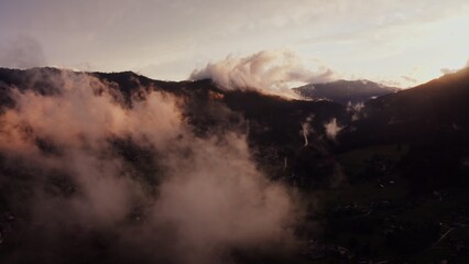 Panoramic view of the cottage town, located in a lowland between the Alpine mountains, the tops of which are covered with white clouds. Alps, Austria, drone video