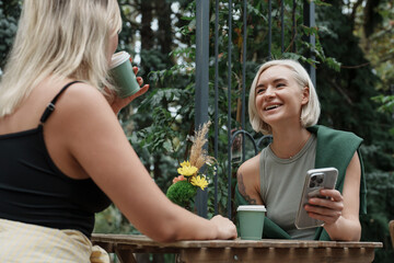 Two friends are enjoying a cheerful conversation over coffee at an outdoor table surrounded by nature. The relaxed atmosphere highlights friendship, communication, and leisure time.