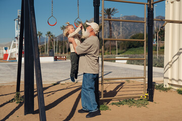 A grandparent and grandchild share a joyful moment at the playground. The child, holding onto the...