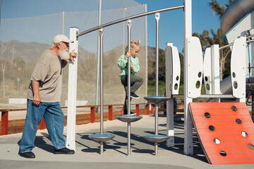 A grandfather stands by, supporting his grandchild as they climb a playground structure,...