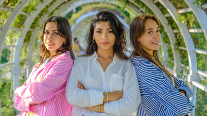 Three young female friends in a tree-filled park on a spring day, smiling as they look at the...