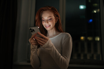 Portrait of happy young woman browsing social media on smartphone, enjoying cozy evening at home, soft screen light illuminating smiling face in tranquil bedroom atmosphere on background of window.