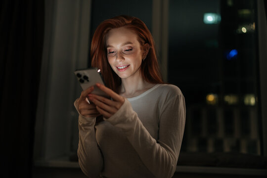 Portrait of happy young woman browsing social media on smartphone, enjoying cozy evening at home, soft screen light illuminating smiling face in tranquil bedroom atmosphere on background of window.