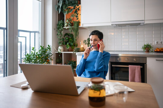 Thoughtful focused mature woman talks on smartphone with pen in mouth, delegating tasks to employees successfully from home. Working on laptop in morning after tea, managing timetable and chats
