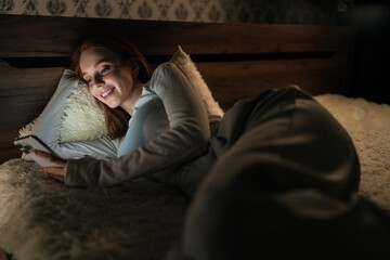 Charming redhead female lying in bed at night using smartphone smiling looking to screen, enjoying leisure time in cozy bedroom, illuminated by phone screen. Concept of mobile addiction and insomnia