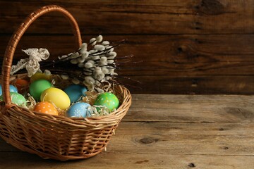 Beautiful decorated Easter eggs in wicker basket and willow branches on wooden table, space for text
