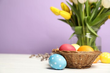 Easter eggs in wicker basket and beautiful tulips on white wooden table against lilac background, closeup. Space for text