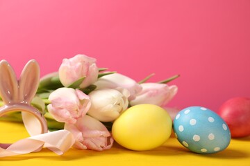 Easter eggs, beautiful tulips and bunny figure on yellow wooden table against pink background, closeup