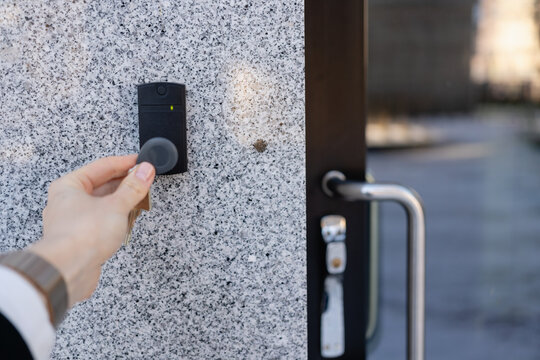 Close-up of a person's hand holding a keychain with a key fob near a key reader access device on a granite building&rsquo;s exterior wall next to glass door, showcasing security and smart access technology