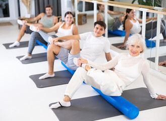 Obraz premium Gracile old-aged woman doing pilates exercises with roller on gray mat during workout session