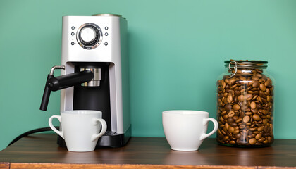 Obraz premium Modern coffee machine, cups and jar with beans on table near green wall. with white shades