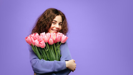 A young woman with curly brown hair and a joyful smile embraces a bouquet of pink tulips against a vibrant purple background. She wears a cozy lavender sweater, radiating warmth and happiness.