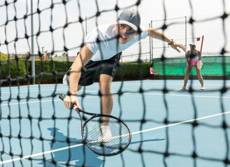 Portrait of emotional determined young guy playing tennis on open court in summer, swinging racket to return ball over net. Sportsman ready to hit volley.