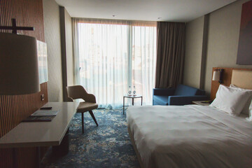 A modern hotel room with a large bed, a blue sofa, a desk, and a floor-to-ceiling window with sheer curtains, allowing soft daylight to enter. The interior is stylish and comfortable