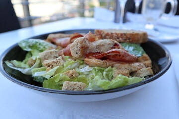 Salad on a black plate consisting of bacon, chicken, lettuce and bread crumbs on a white tablecloth background