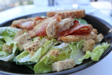 Salad on a black plate consisting of bacon, chicken, lettuce and bread crumbs on a white tablecloth background