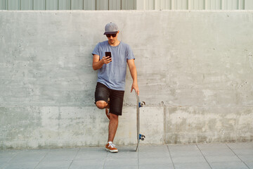 Young skateboarder using smartphone leaning against concrete wall